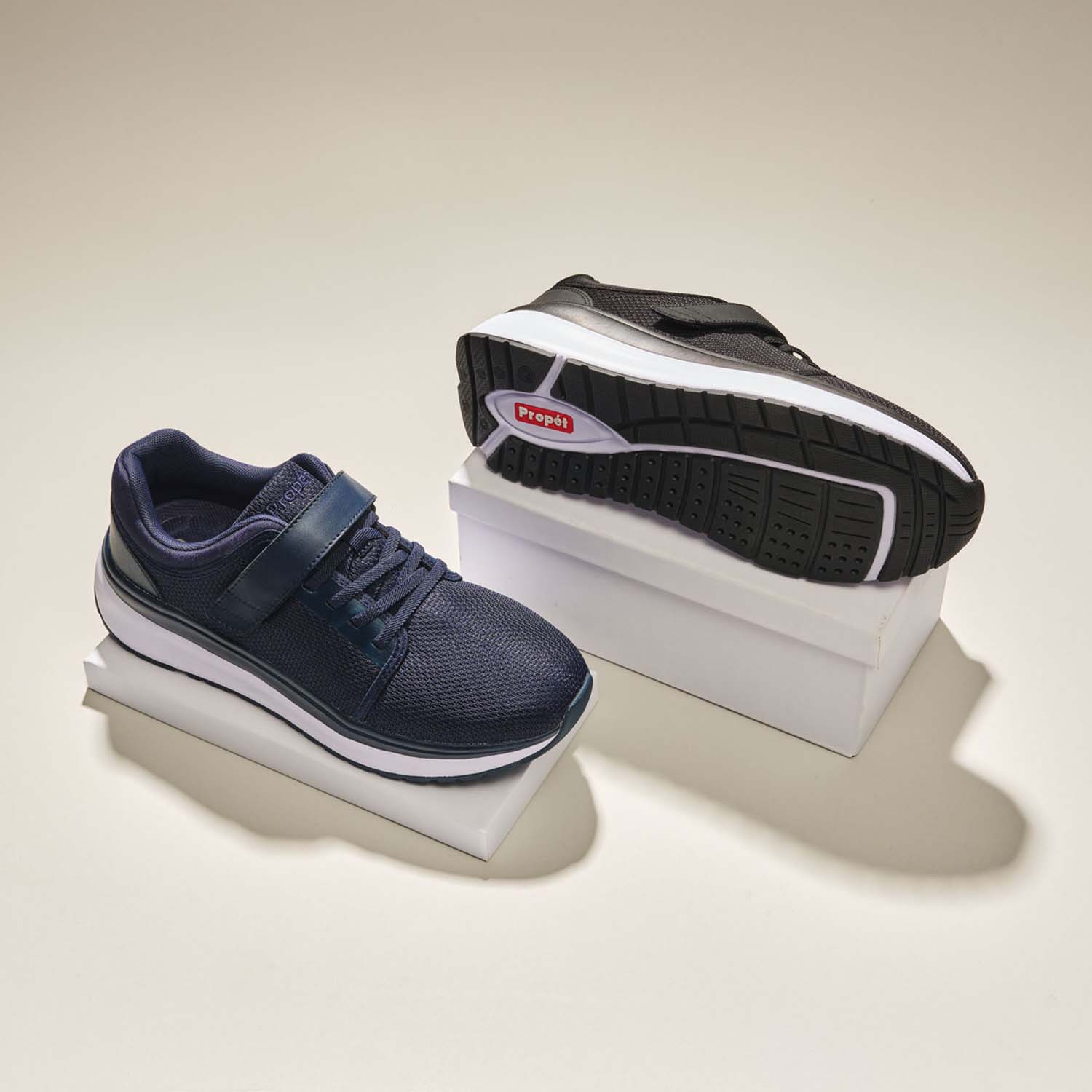 Angled, top down view of the two color options for the Ultima FX shoes (Black & Navy) side-by-side on separate platforms with beige backdrop